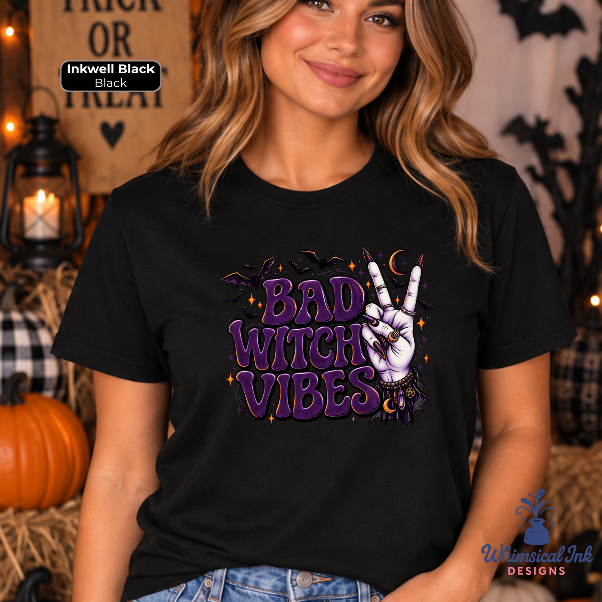 A woman wearing a black graphic t-shirt with a purple and white 'Bad Witch Vibes' design, posed in a Halloween-themed setting with pumpkins and a lantern.