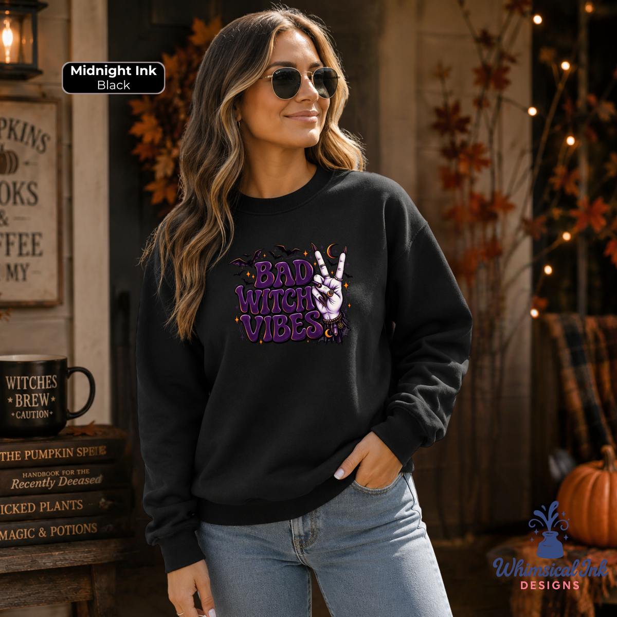 A woman wearing a black crewneck sweatshirt with a purple and white graphic that reads 'BAD WITCH VIBES', paired with light blue jeans in a cozy autumn-themed setting.