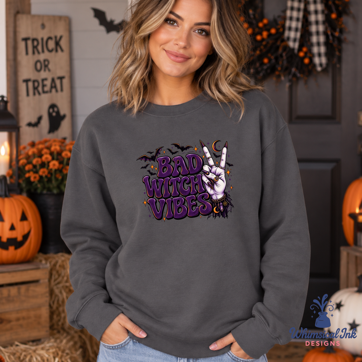 A woman wearing a dark gray crewneck sweatshirt with a 'Bad Witch Vibes' graphic on the front, posed in a cozy, decorated Halloween-themed room.