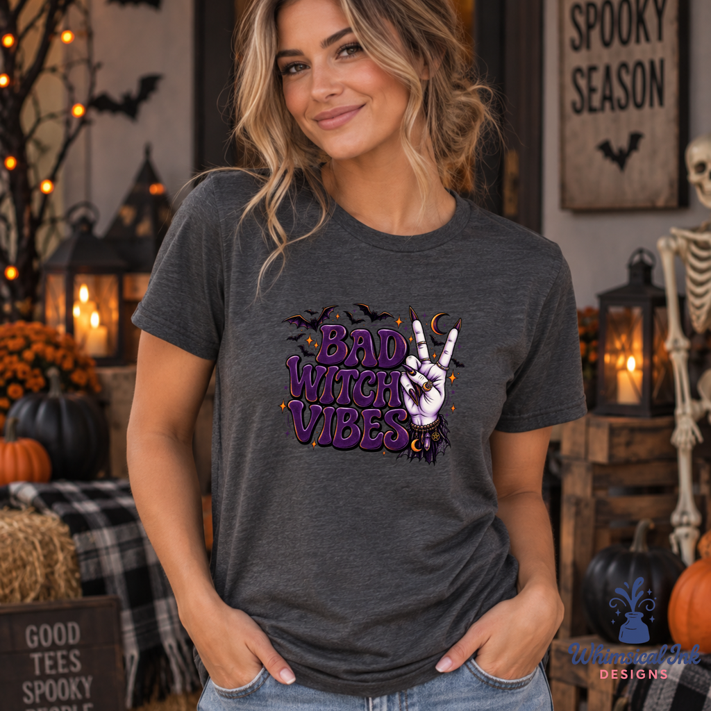 A woman wearing a dark heather gray graphic t-shirt with a purple and white 'Bad Witch Vibes' design, posed in a cozy, decorated Halloween-themed room with pumpkins, lanterns, and a skeleton.