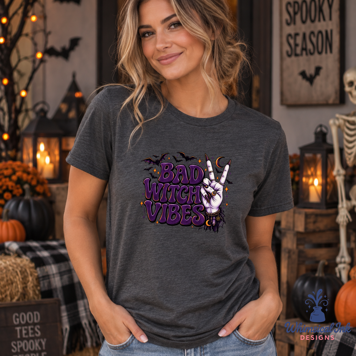 A woman wearing a dark heather gray graphic t-shirt with a purple and white 'Bad Witch Vibes' design, posed in a cozy, decorated Halloween-themed room with pumpkins, lanterns, and a skeleton.