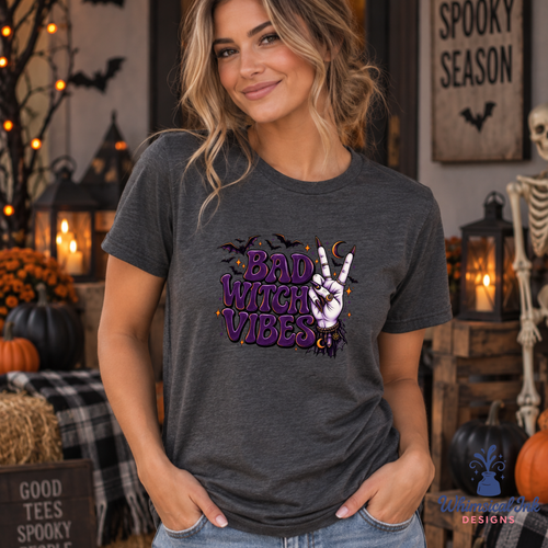 A woman wearing a dark heather gray graphic t-shirt with a purple and white 'Bad Witch Vibes' design, posed in a cozy, decorated Halloween-themed room with pumpkins, lanterns, and a skeleton.