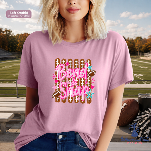 A woman wearing a light pink graphic t-shirt with a 'Bend & Snap' football theme, set against an outdoor football field background.