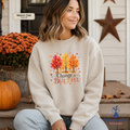 A woman wearing a sand-colored sweatshirt with a colorful autumn tree graphic and the text 'Change is Beautiful' sitting on a porch. The sweatshirt has a color label in the top left that reads 'Warm Oat Sand' and a brand logo in the bottom right that reads Whimsical Ink Designs'.