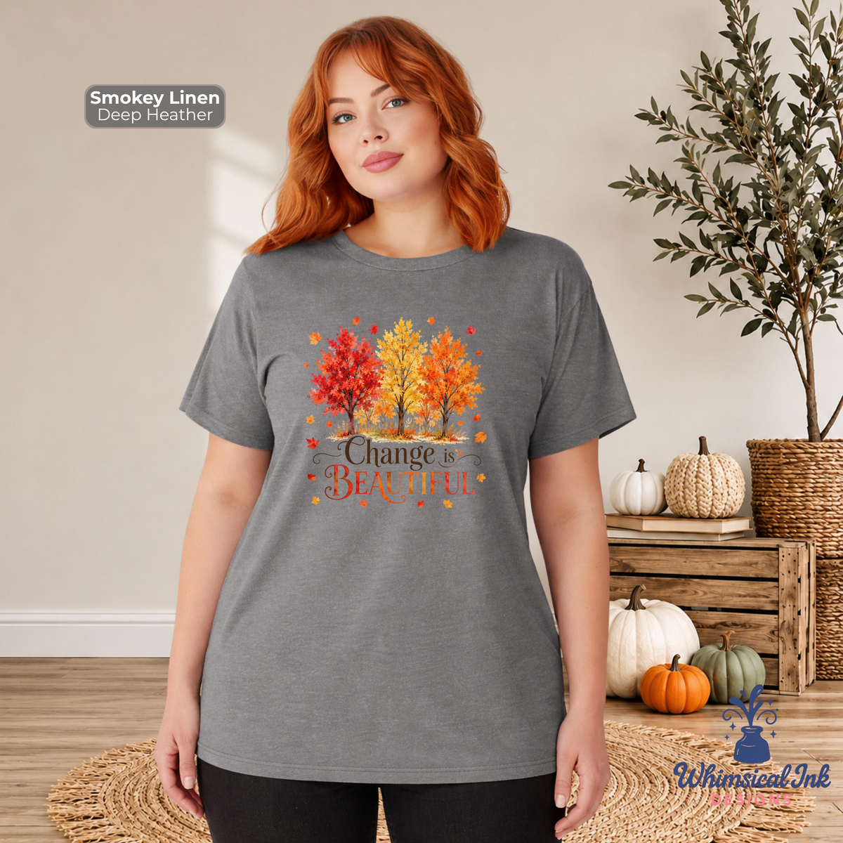 A woman modeling a heather gray t-shirt with a colorful autumn tree graphic and the text 'Change is Beautiful'. The background is a cozy, styled room with fall decorations including pumpkins and a potted plant.