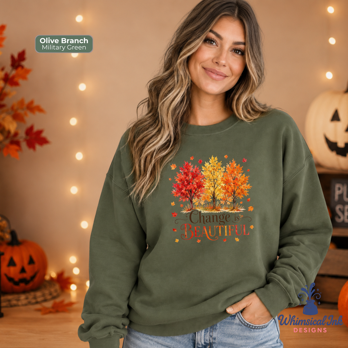 A woman wearing an olive branch sweatshirt with a colorful autumn tree graphic and the text 'Change is Beautiful' in a cozy, Halloween-themed setting with pumpkins and string lights.