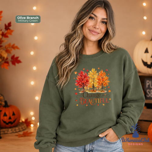 A woman wearing an olive branch sweatshirt with a colorful autumn tree graphic and the text 'Change is Beautiful' in a cozy, Halloween-themed setting with pumpkins and string lights.