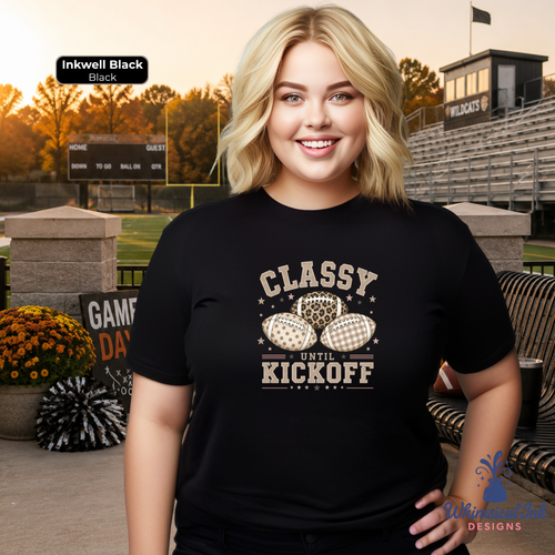 A woman stands outdoors at a stadium wearing a black graphic T-shirt with a distressed cream print that reads “CLASSY UNTIL KICKOFF” above two footballs. The scene includes bleachers, a scoreboard, flowers, and a seating area, with a small “Inkwell Black” label in the top left and a “WildCats” sign in the background.