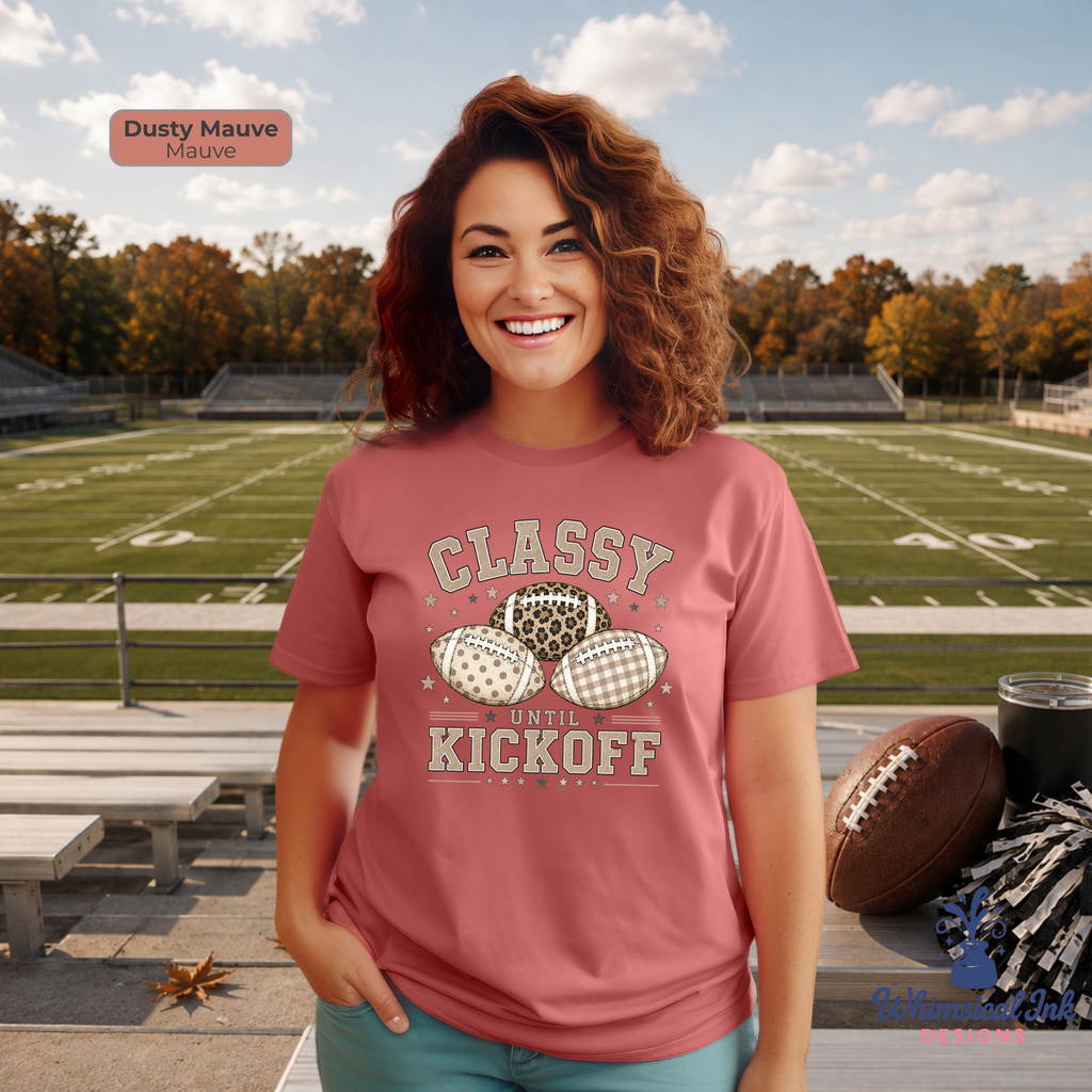 A woman stands on a football field wearing a dusty mauve graphic t-shirt with a vintage-style football print that reads 'CLASSY KICKOFF'. The image includes a color swatch label in the top left indicating 'Dusty Mauve (Mauve)' and a brand watermark in the bottom right corner.