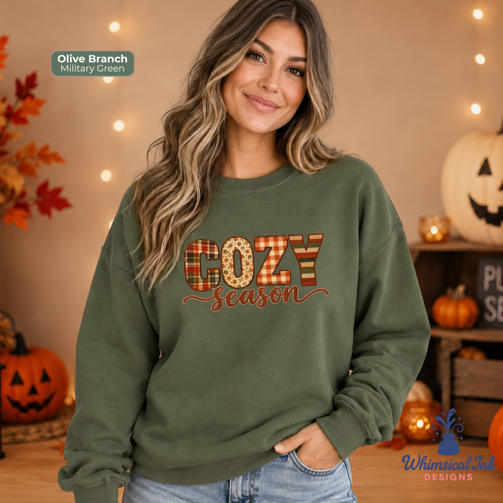 A woman wearing an olive green sweatshirt with a 'COZY Season' graphic in a fall-themed setting with pumpkins and string lights. The graphic features the word 'COZY' in a plaid pattern and 'Season' in cursive script.