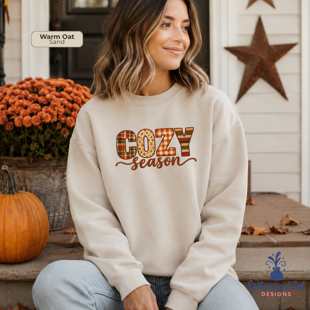 A woman sits on porch steps wearing a cream sweatshirt with a fall-themed 'COZY Season' graphic. The graphic features 'COZY' in a plaid pattern of orange, brown, and beige, with 'Season' written below in brown cursive.