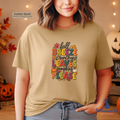 A woman wearing a Golden Sand heather sand dune graphic t-shirt with a colorful autumn-themed text print that reads 'fall Breeze Crunchy LEAVES pumpkins PLEASE'. The background depicts a cozy, warm fall setting with string lights, pumpkins, and autumn leaves.