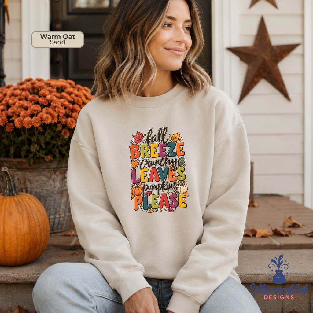 A woman sits on a porch wearing a cream-colored crewneck sweatshirt with a colorful autumn-themed graphic that reads 'all BREEZE crunchy LEAVES are PLEASE' surrounded by fall leaves and pumpkins. The background shows a white shiplap wall, a rustic star, orange mums, and a pumpkin.