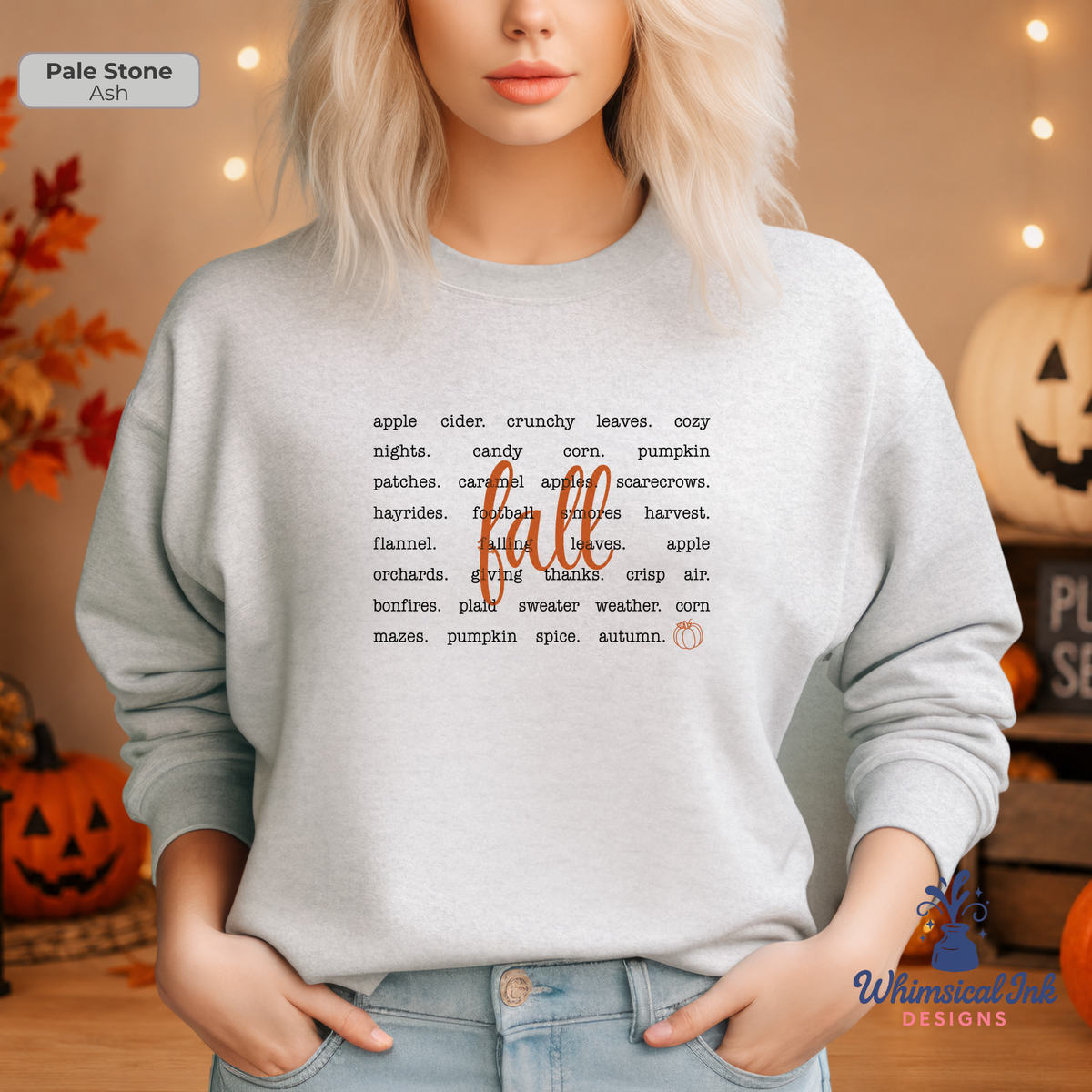 A woman wearing a pale ash crewneck sweatshirt with a 'fall' word graphic printed in black and orange, posed in a cozy, warmly lit autumn-themed room with pumpkins and string lights.