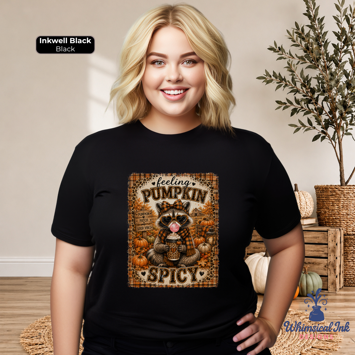 A smiling blonde woman models a black short-sleeve graphic T-shirt in a warm indoor setting with pumpkins and a plant. The shirt features a central autumn-themed print with the words 'feeling PUMPKIN SPICY' and an illustrated raccoon holding a coffee cup.