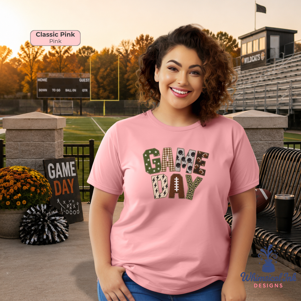 A woman stands on a sports field at sunset wearing a pink graphic T-shirt that reads 'GAME DAY' in football-themed lettering. The background includes bleachers, a scoreboard, flowers, and a bench.
