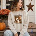 A woman wearing a cream-colored crewneck sweatshirt with a Halloween-themed graphic of a ghost in a library, sitting on porch steps next to a pumpkin and orange flowers.
