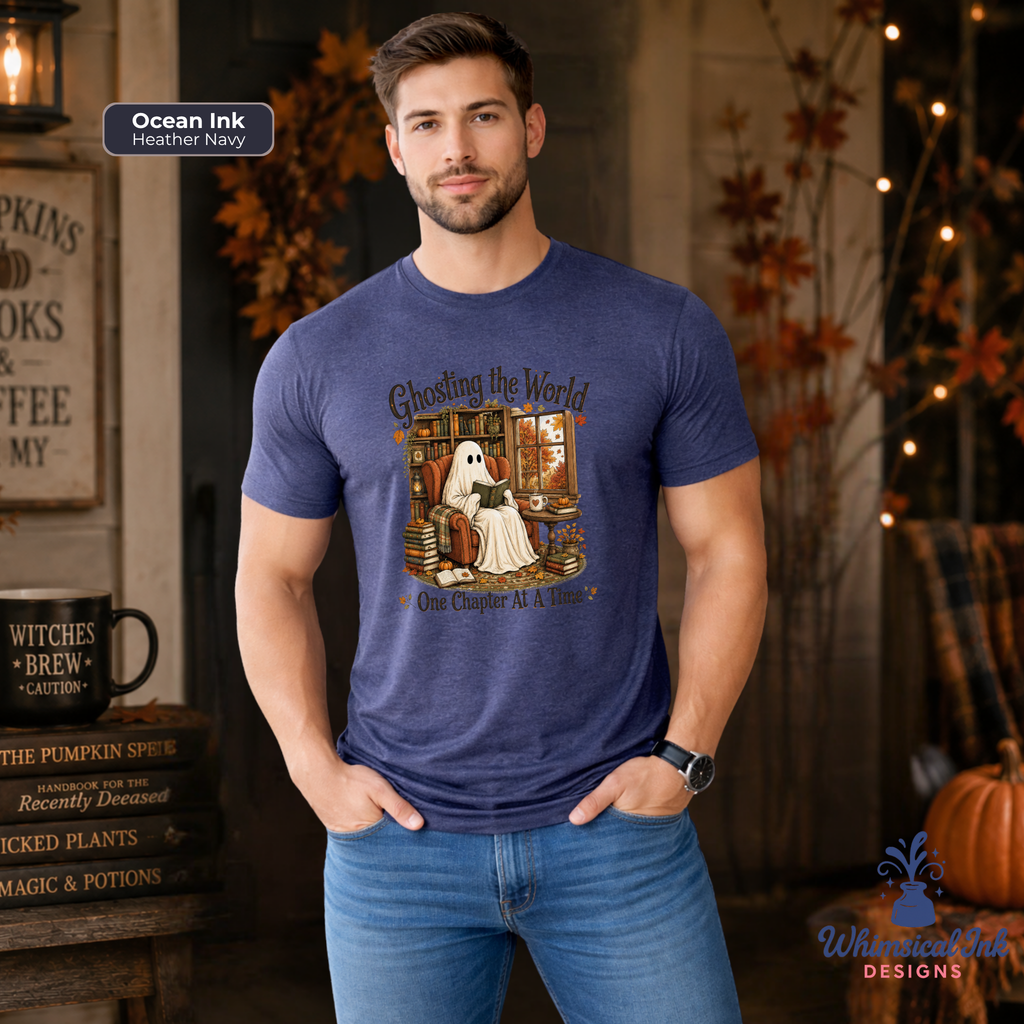 A man wearing a heather navy blue graphic t-shirt with a Halloween-themed ghost illustration and the text 'Ghosting the World One Chapter At A Time'. The background is a cozy, warmly lit autumn-themed room with string lights, pumpkins, and rustic decor.