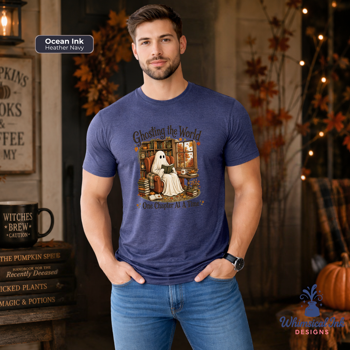 A man wearing a heather navy blue graphic t-shirt with a Halloween-themed ghost illustration and the text 'Ghosting the World One Chapter At A Time'. The background is a cozy, warmly lit autumn-themed room with string lights, pumpkins, and rustic decor.