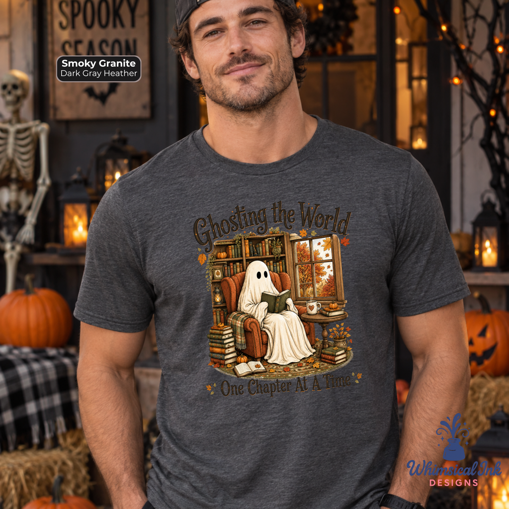 A man wearing a dark heather gray graphic t-shirt with a Halloween ghost illustration and the text 'Ghosting the World One Chapter At A Time'. The background is a cozy, warmly lit Halloween-themed room with pumpkins, a skeleton, and string lights.