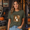 A woman wearing a military green graphic t-shirt with a ghost-themed illustration and the text 'Ghosting the World One Chapter At A Time'. The background is a cozy, decorated Halloween scene with pumpkins, a skeleton, and warm lighting.