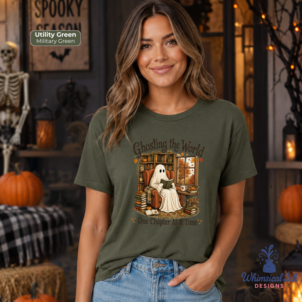 A woman wearing a military green graphic t-shirt with a ghost-themed illustration and the text 'Ghosting the World One Chapter At A Time'. The background is a cozy, decorated Halloween scene with pumpkins, a skeleton, and warm lighting.