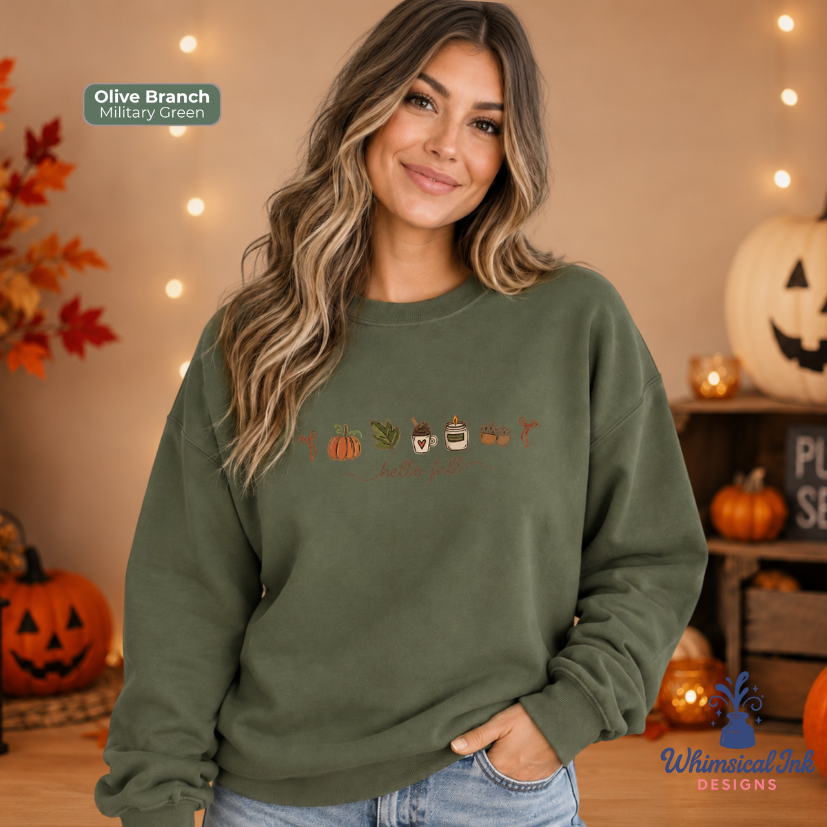 A woman wearing an olive branch military green crewneck sweatshirt with a 'Thanksgiving' graphic featuring pumpkins, turkeys, and a turkey foot. The background is a cozy, warm autumn-themed setting with string lights, pumpkins, and fall foliage.