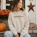 A woman sitting on porch steps wearing a cream-colored crewneck sweatshirt with a fall-themed graphic that reads 'hello fall' with small autumn icons. The image has a 'Warm Oat Sand' color label overlay and a 'Whimsical Ink DESIGNS' logo in the bottom right corner.