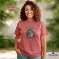 Woman wearing a dusty mauve t-shirt with a graphic design and text, standing outdoors.