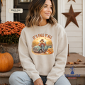 A woman wearing a cream-colored crewneck sweatshirt with a fall-themed graphic that reads 'IT'S FALL Y'ALL' and shows a cartoon character in a cowboy hat leaning on a vintage truck next to pumpkins. She is sitting on a porch with a pumpkin and orange flowers in the background.