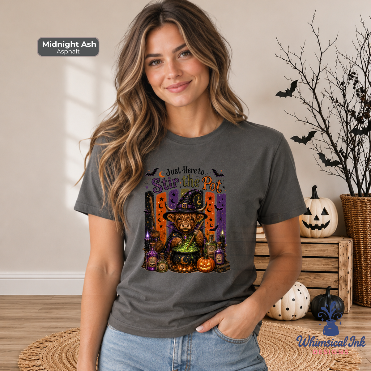 A woman wearing a dark gray graphic t-shirt with a Halloween-themed design featuring a highland cow dressed like a witch and pumpkins. The t-shirt is labeled 'Midnight Ash Asphalt' in a badge in the top left corner, and the brand 'Whimsical Ink' is visible as a watermark in the bottom right corner.