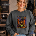 A woman wearing a dark gray crewneck sweatshirt with a colorful graphic of a witch cat stirring a cauldron. The background is a cozy, Halloween-themed room with a cauldron, broom, and books.