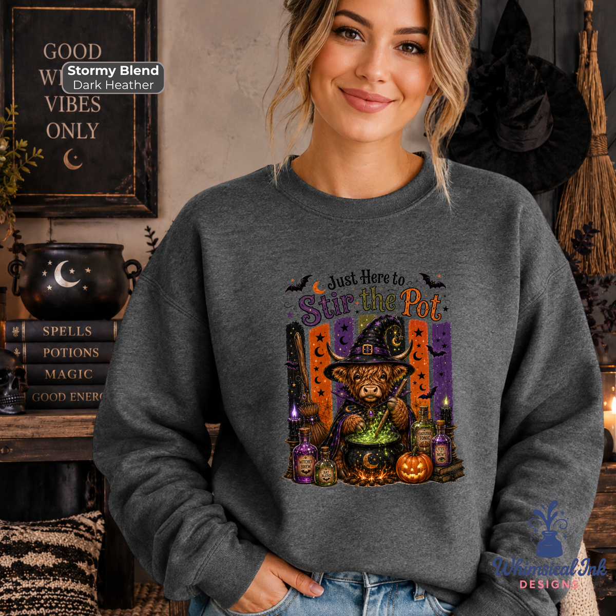 A woman wearing a dark gray crewneck sweatshirt with a colorful graphic of a witch cat stirring a cauldron. The background is a cozy, Halloween-themed room with a cauldron, broom, and books.