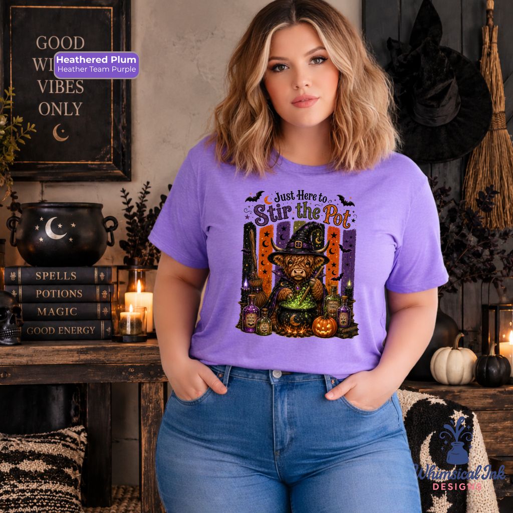 A woman wearing a lavender-purple graphic t-shirt with a 'Just Here to Stir the Pot' witch design, paired with blue jeans, standing in a cozy, dark-themed Halloween room with candles, a cauldron, and pumpkins.