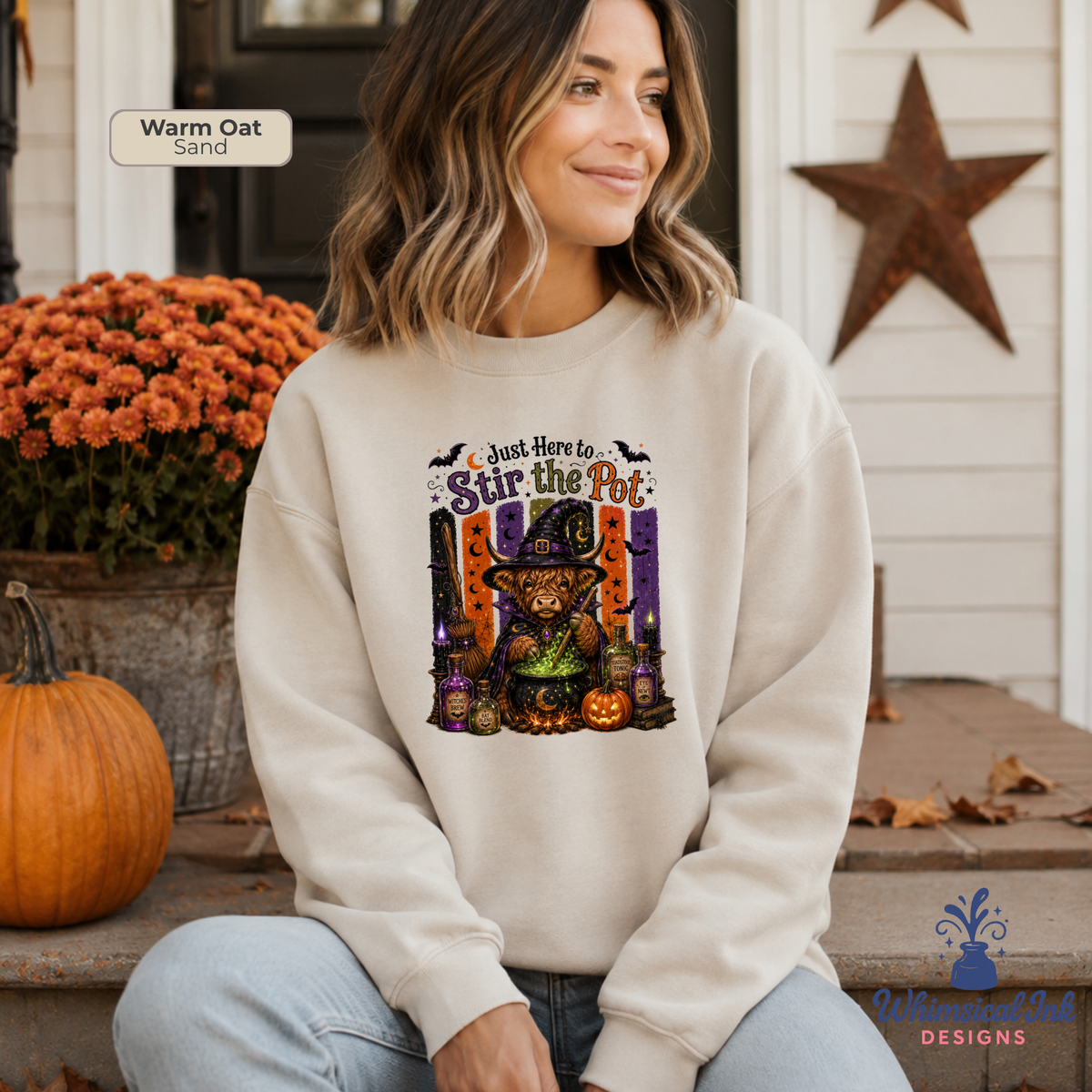 A woman sits on a porch wearing a cream sweatshirt with a colorful graphic of a witch cat in a hat and the text 'Just Here to Stir the Pot'. The image includes a color swatch label reading 'Warm Oak Sand' and a brand logo for 'Whimsical Ink Designs' in the bottom right corner.