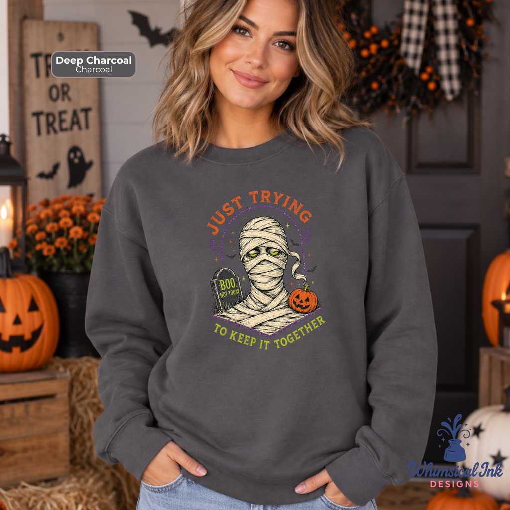 A woman wearing a dark charcoal gray sweatshirt with a Halloween graphic of a mummy and the text 'JUST TRYING TO KEEP IT TOGETHER'. The background is a cozy, decorated Halloween scene with jack-o'-lanterns, pumpkins, and a 'TRICK OR TREAT' sign.