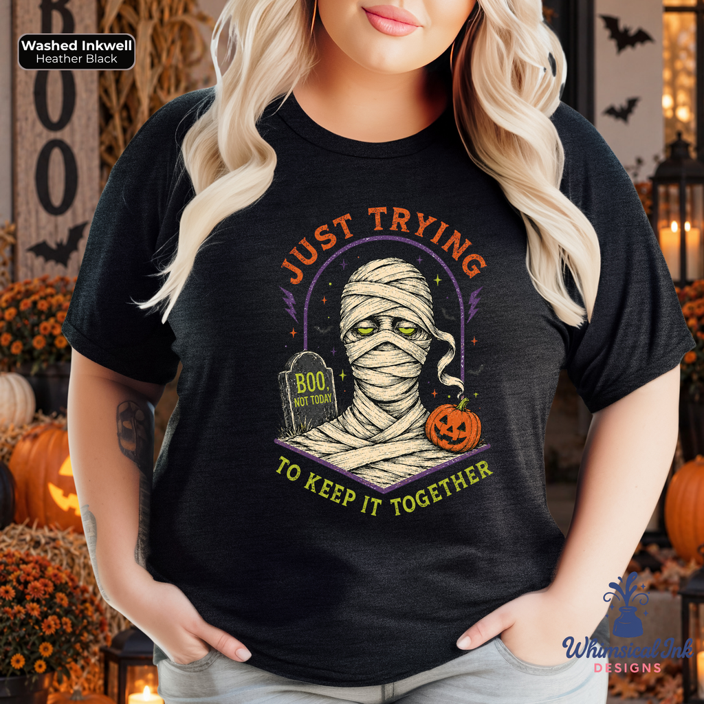 A woman wearing a black graphic t-shirt with a Halloween-themed design of a bandaged mummy holding a pumpkin next to a tombstone that reads 'Just Trying to Keep It Together'. The background is a decorated autumn scene with pumpkins and candles.