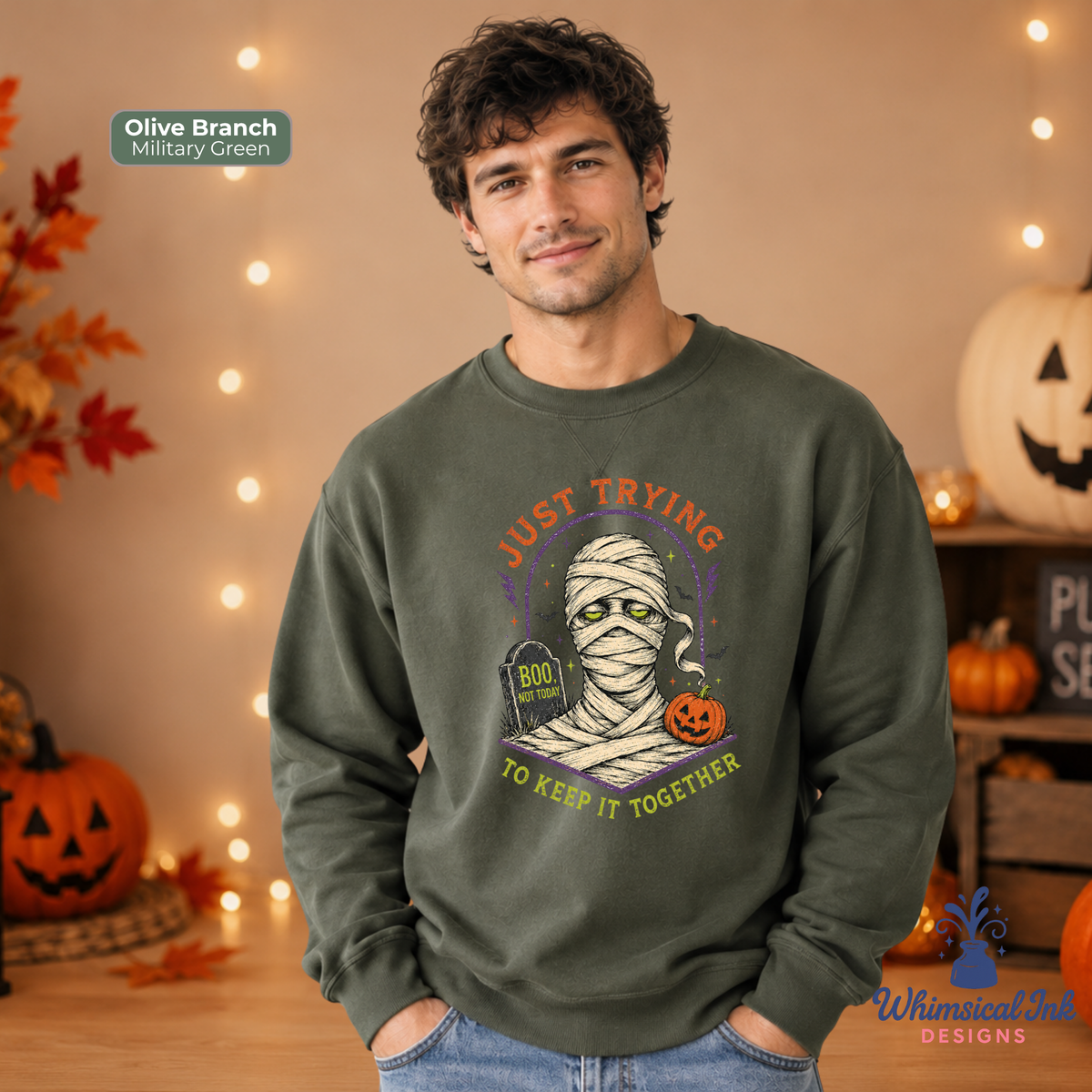 A man wearing a military green crewneck sweatshirt with a Halloween graphic of a mummy with a tombstone that says 'BOO NOT TODAY' and the text 'JUST TRYING TO KEEP IT TOGETHER'. The background is a warm, cozy fall setting with string lights, pumpkins, and autumn leaves.