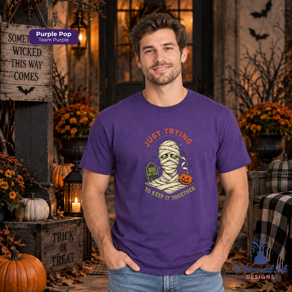 A man wearing a purple graphic t-shirt with a Halloween mummy design, standing in a decorated autumn-themed setting with pumpkins and fall foliage.