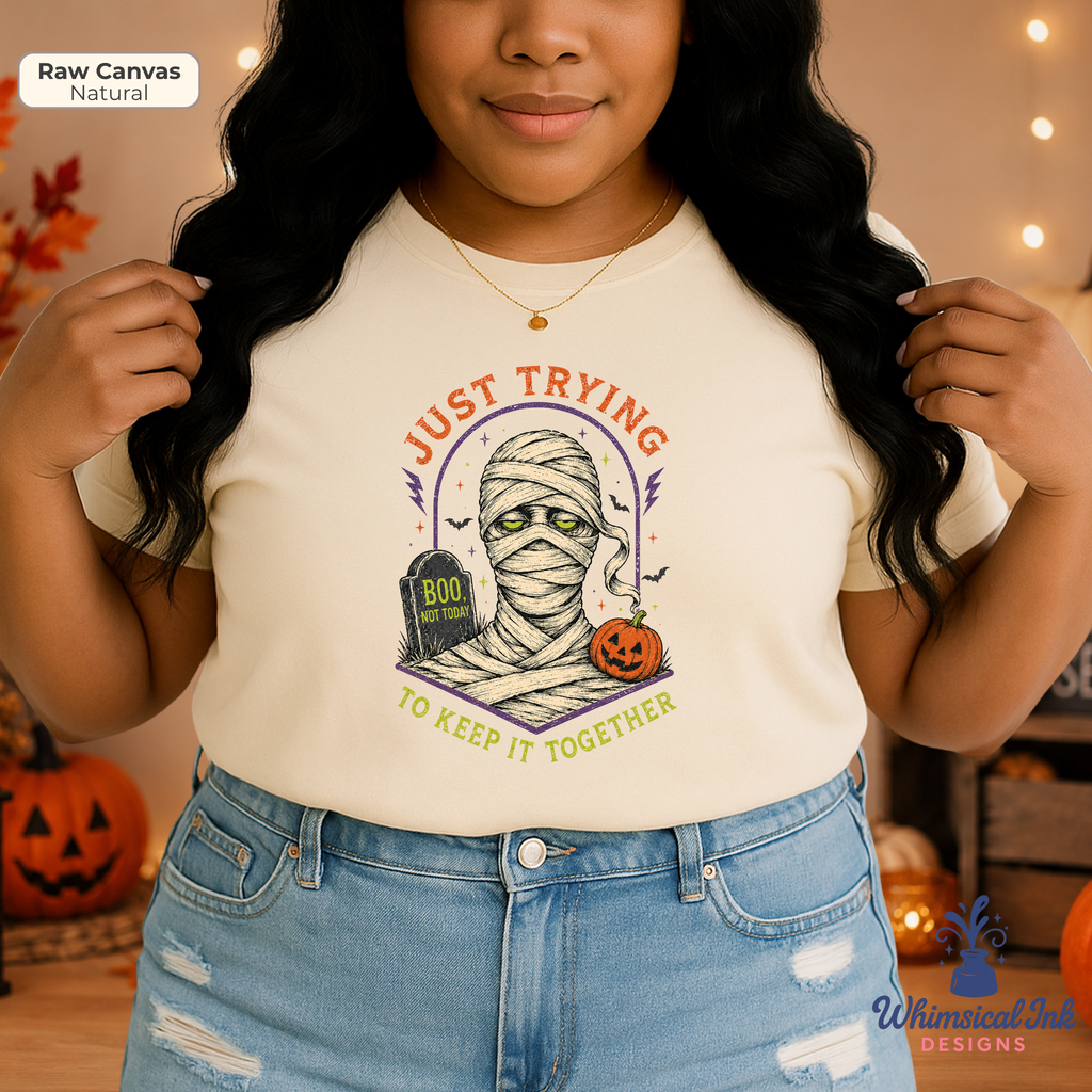 A woman wearing a cream-colored graphic t-shirt with a Halloween mummy design and the text 'JUST TRYING TO KEEP IT TOGETHER'. She is wearing light-wash distressed denim jeans, standing in a cozy, warmly lit room with pumpkins and string lights in the background.