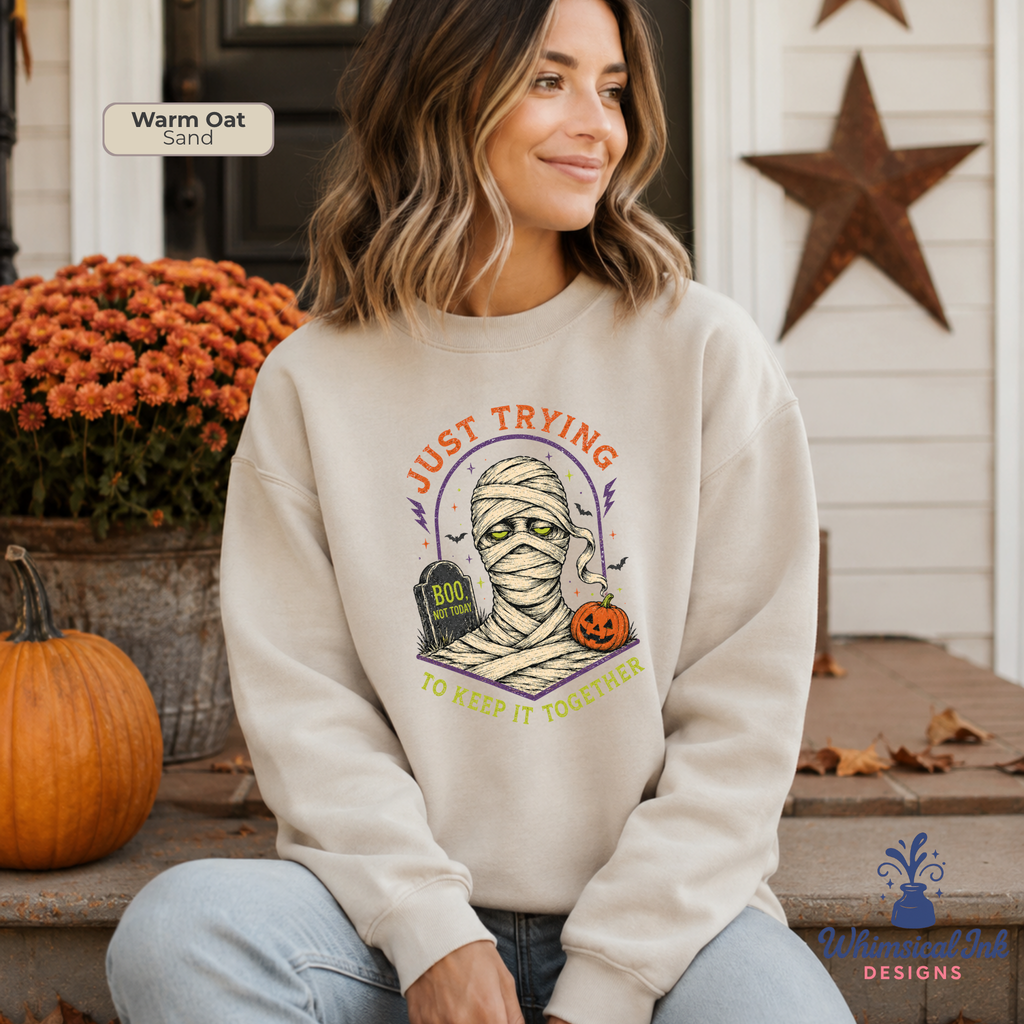 A woman sits on porch steps wearing a cream-colored sweatshirt with a Halloween graphic that reads 'JUST TRYING TO KEEP IT TOGETHER' around a mummy and tombstone design. The image has a 'Warm Oat Sand' color label in the top left, and a Whimsical Ink Designs logo.