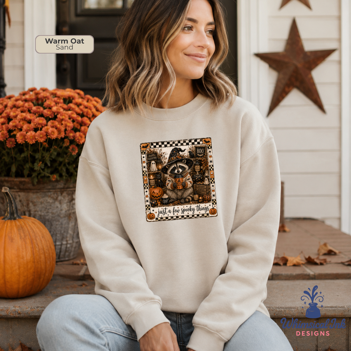 A woman wearing a cream-colored sweatshirt with a Halloween-themed graphic of a raccoon holding a lantern and the text 'just a few spooky things' at the bottom. The scene is set on a porch with pumpkins, orange flowers, and a rustic star decoration.