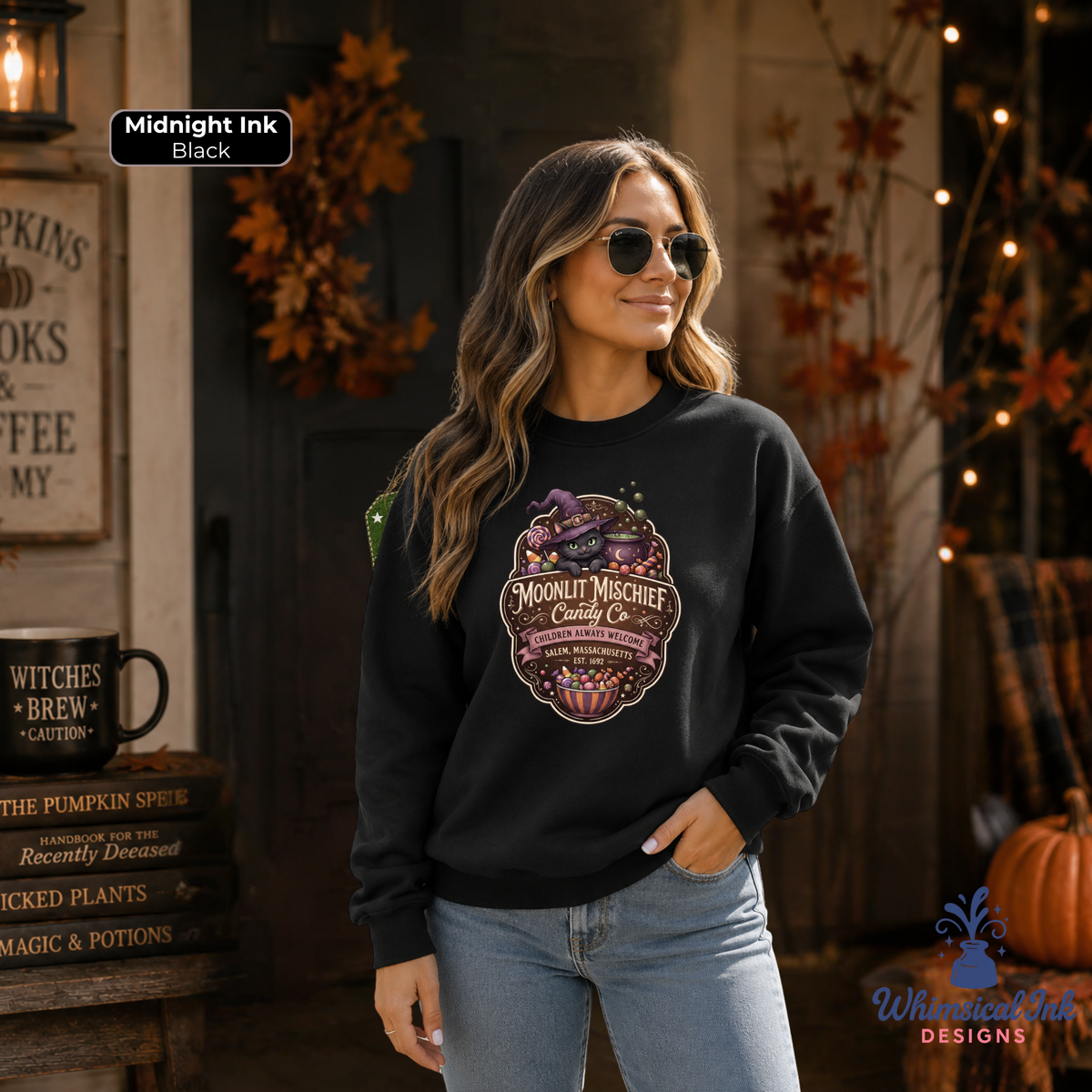 A woman wearing a black crewneck sweatshirt with a large Halloween-themed graphic that reads 'Mortally Weird Candy Co.' in a spooky autumn setting with fall leaves and pumpkins.