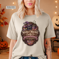 A woman wearing a light heathered t-shirt with a Halloween graphic of a black cat in a witch hat next to a cauldron of candy. The text on the shirt reads 'Moonlit Mischief Candy Co'.
