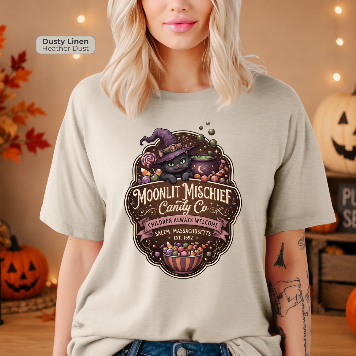 A woman wearing a light heathered t-shirt with a Halloween graphic of a black cat in a witch hat next to a cauldron of candy. The text on the shirt reads 'Moonlit Mischief Candy Co'.