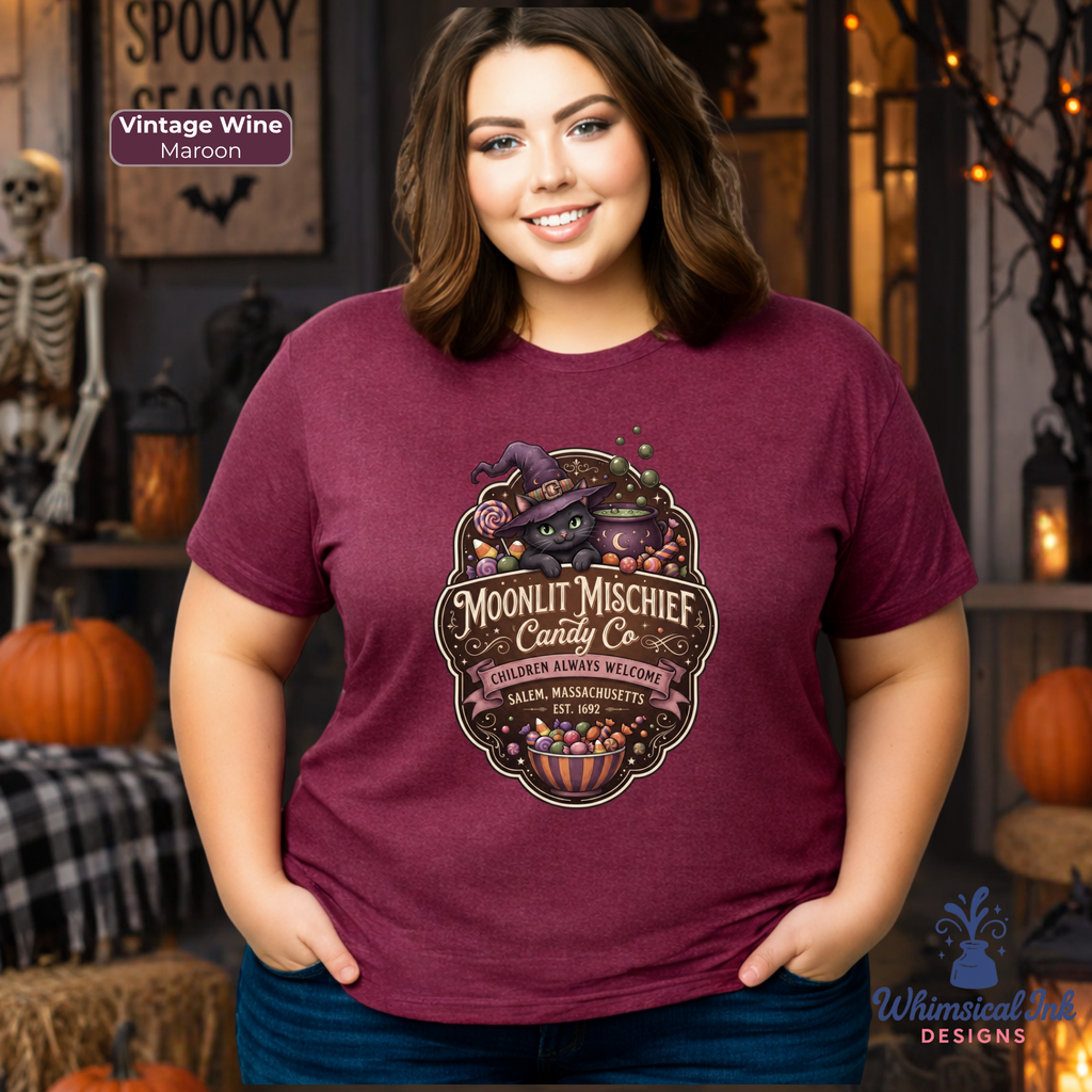 A woman wearing a maroon graphic t-shirt with a Halloween-themed 'Moonlit Mischief Candy Co.' design, posed in a cozy, decorated Halloween-themed room with pumpkins and a skeleton.