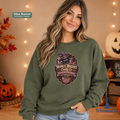 A woman wearing an olive green sweatshirt with a Halloween-themed graphic that reads 'Moonlit Mischief Candy Co.' She is posed in a cozy, warmly lit fall setting with pumpkins and string lights.