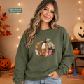 A woman wearing an olive branch sweatshirt with a patchwork pumpkin design on the front, posed in a cozy autumn-themed room with string lights and pumpkins.