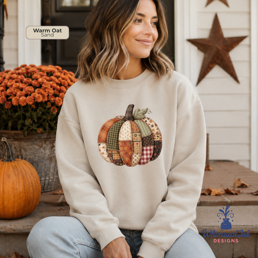 A woman wearing a cream-colored sweatshirt with a patchwork pumpkin graphic sits on a porch. The sweatshirt has a warm oatmeal/sand base color with a multi-colored autumn-themed pumpkin design in the center.