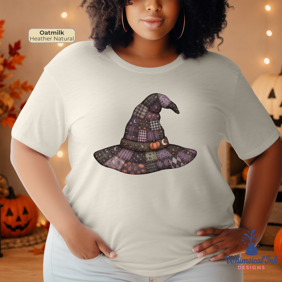 A woman wearing a light oatmeal heather natural t-shirt with a purple patchwork witch hat graphic on the front, posed in a cozy, warm autumn-themed room with pumpkins and string lights.
